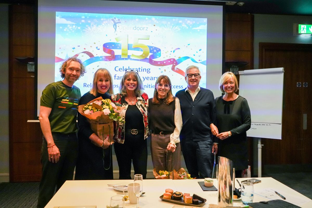 A group of six people smiling and posing indoors with a colorful presentation slide behind them celebrating fifteen years. Two hold bouquets, conveying joy.
