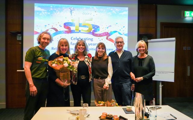 A group of six people smiling and posing indoors with a colorful presentation slide behind them celebrating fifteen years. Two hold bouquets, conveying joy.